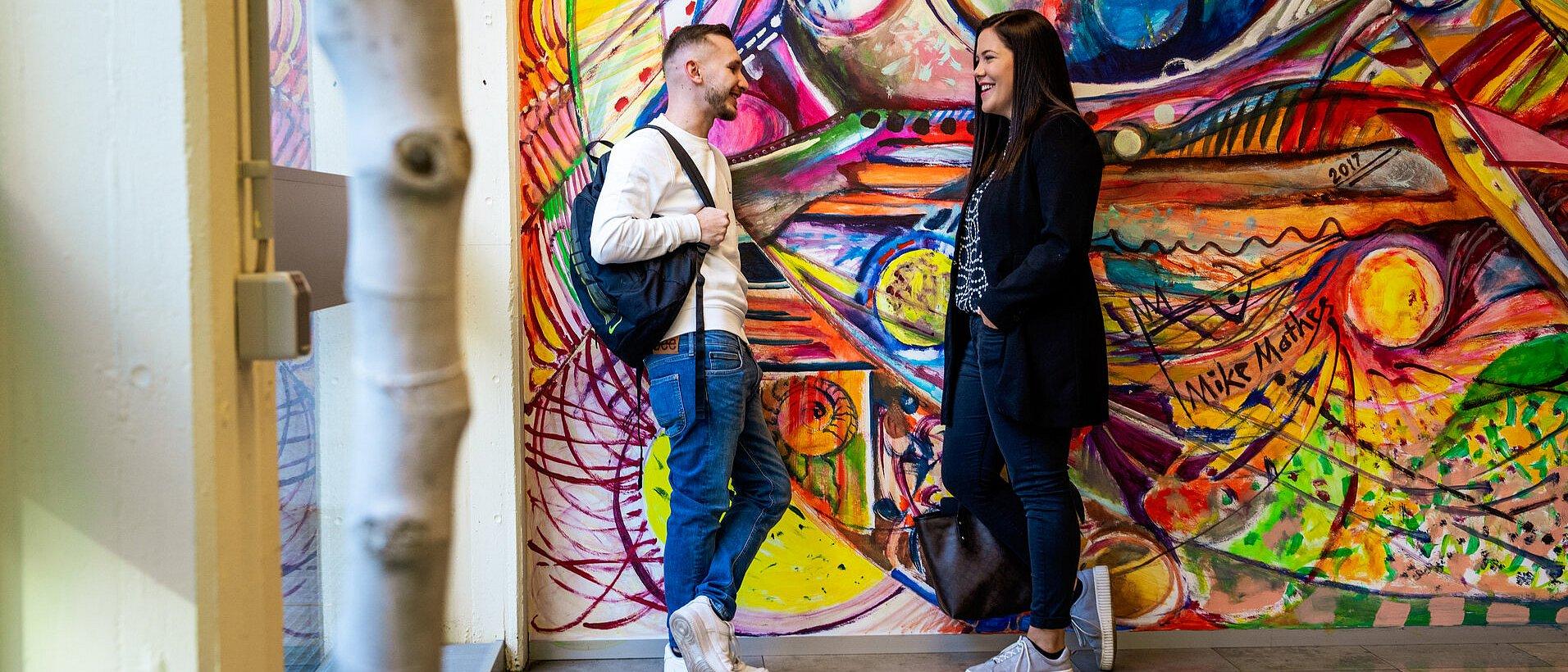 Zwei junge Erwachsene stehen vor einer bunten Wandmalerei und unterhalten sich.