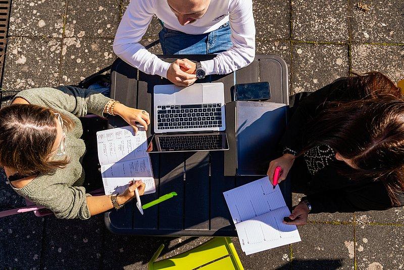 Draufsicht: Drei Personen arbeiten draußen an einem Tisch mit Laptop und Heften.