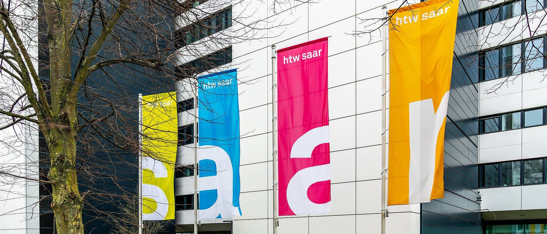 Vier bunte Fahnen mit „htw saar“ vor einem Gebäude.