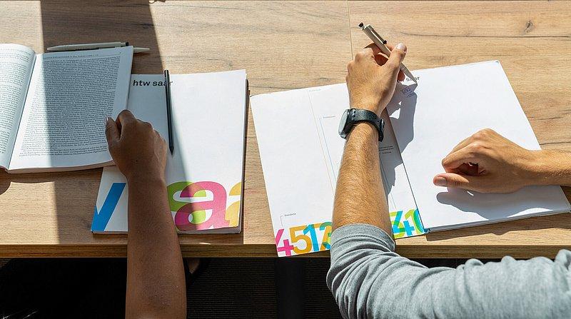 Zwei Personen arbeiten an Schreibtisch mit Büchern und Blättern in der Sonne.