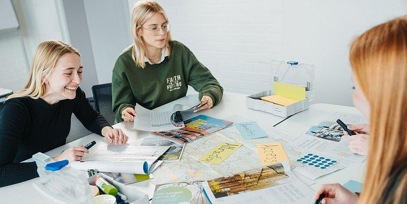 Drei junge Frauen blättern in Magazinen an einem Tisch mit Karten und Notizen.