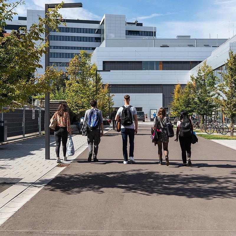 Fünf Personen mit Rucksäcken gehen auf ein modernes Hochschulgebäude zu.