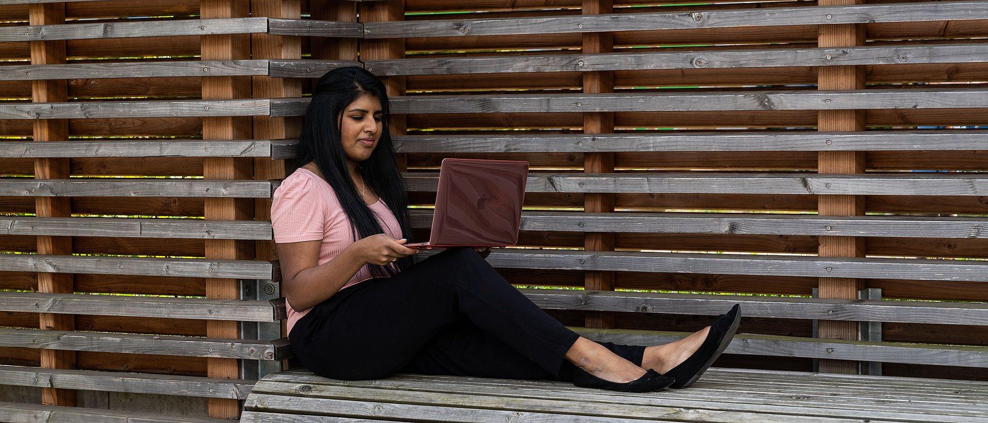 Person sitzt auf einer Holzbank und arbeitet mit einem Laptop.