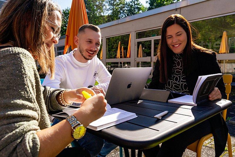 Drei junge Erwachsene sitzen draußen am Tisch mit Laptop, Buch und Notizen.