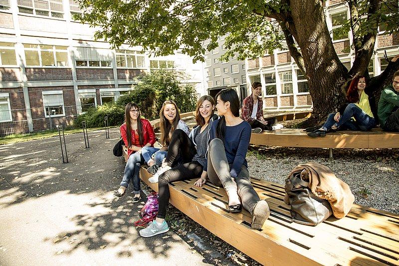 Mehrere Personen sitzen lachend auf Bänken unter einem Baum im Hof.