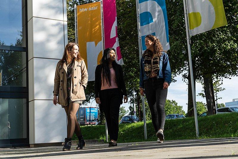 Drei Personen gehen draußen an Fahnen mit der Aufschrift „htw saar“ vorbei.
